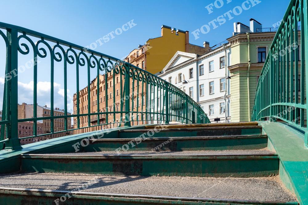 На фотографии изображен Сенной мост через канал Грибоедова в Санкт-Петербурге. Вдали видны здания исторического центра Санкт-Петербурга с их характерными фасадами. Яркое солнечное освещение добавляет тепла и оживляет этот уголок города. Сенной мост — важная часть инфраструктуры Санкт-Петербурга и живописное место для прогулок вдоль канала Грибоедова.